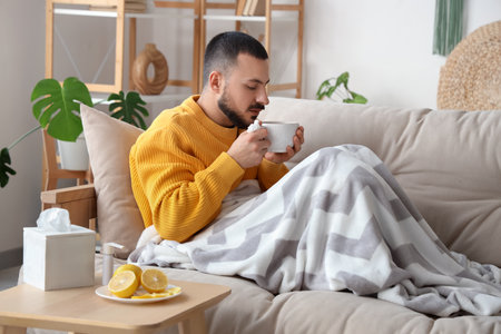 Ill young man drinking lemon tea on sofa at homeの写真素材