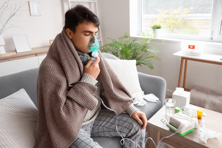 Ill young man with plaid using nebulizer on sofa at homeの写真素材