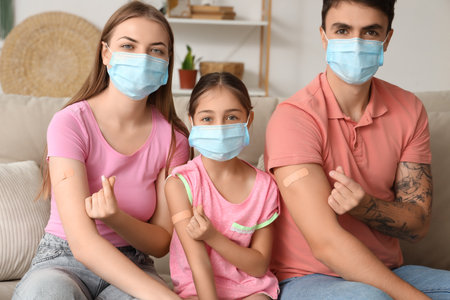 Family in masks with medical patches after vaccination making heart gesture on sofa at homeの写真素材