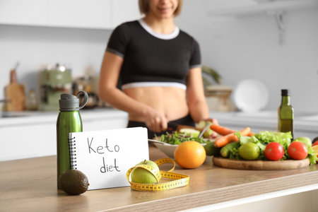 Notebook with text KETO DIET, food and water bottle on table against sporty woman in kitchen, closeupの写真素材