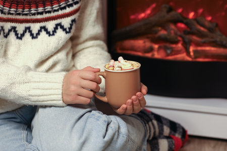 Beautiful young woman with cup of hot chocolate and marshmallows sitting on floor near fireplace at home, closeupの写真素材