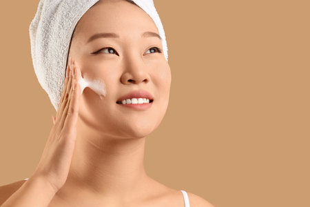 Young Asian woman in towel applying cleansing foam on beige background, closeupの写真素材