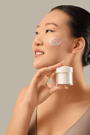 Young Asian woman with jar of facial cream on gray background, closeupの写真素材