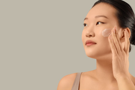 Young Asian woman applying facial cream on gray background, closeupの写真素材