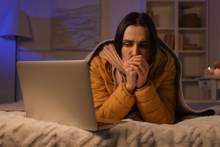 Frozen young woman with laptop lying in bedroom during blackoutの写真素材