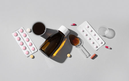 Bottle of cough syrup with measuring cups, spoon and different pills on gray backgroundの写真素材