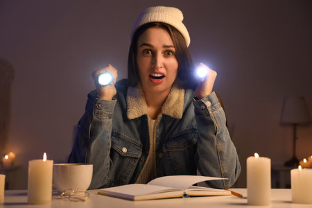 Shocked young woman with flashlights and book on table at home during blackoutの写真素材