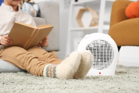 Cute boy in warm socks with electric fan heater reading book on carpet at home, closeupの写真素材