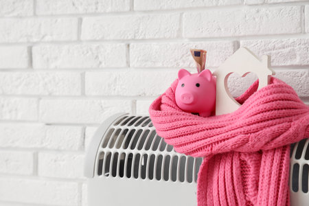 Piggy bank with house model, warm scarf and money on convector heater near white brick wall. Heating saving conceptの写真素材