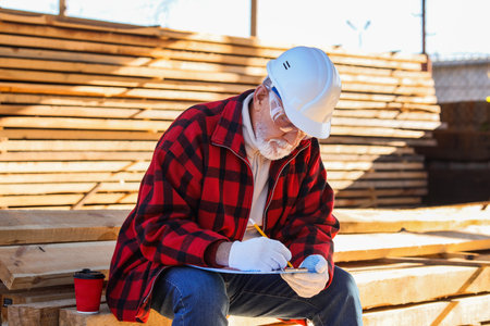 Mature male carpenter with cup of coffee writing in clipboard at sawmillの写真素材