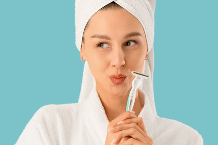Beautiful young thoughtful woman in bathrobe with razor on blue backgroundの写真素材