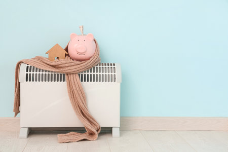 Piggy bank with house model, warm scarf and money on convector heater against blue background. Heating saving conceptの写真素材