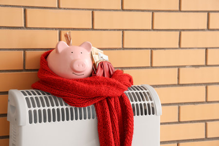Piggy bank with money and warm scarf on convector heater near brick wall. Heating saving conceptの写真素材