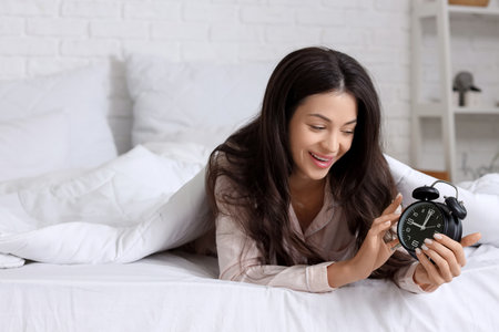 Happy young woman with alarm clock lying under blanket in bedroomの写真素材