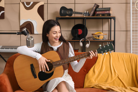 Beautiful young happy female composer with microphone playing guitar on sofa in music studioの写真素材