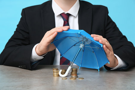 Businessman with toy umbrella and coins at table on blue background, closeupの写真素材