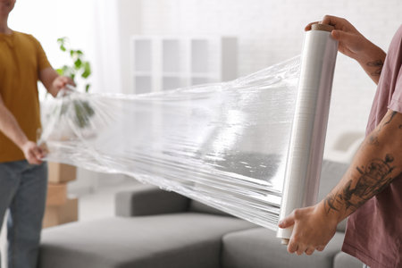 Young men wrapping sofa with stretch film in room on moving dayの写真素材