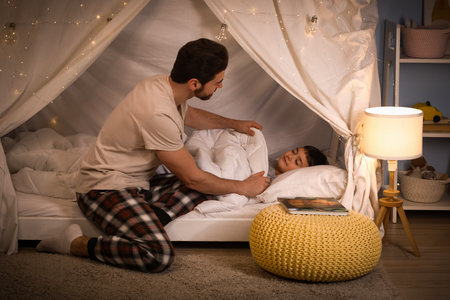 Father covering his sleeping little son with blanket in hut at homeの写真素材