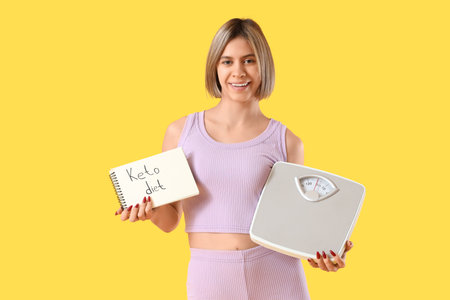Sporty young woman holding notebook with text KETO DIET and weight scales on yellow backgroundの写真素材