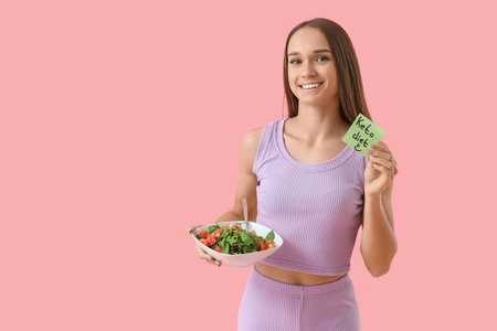 Sporty young woman holding paper with text KETO DIET and vegetable salad on pink backgroundの写真素材