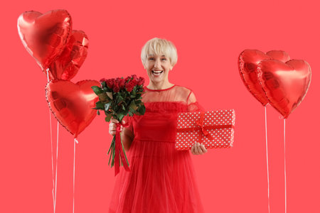 Happy mature woman with gift box, bouquet of roses and heart-shaped balloons on red background. Valentine's Day celebrationの写真素材