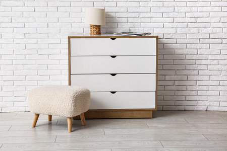 Pouf and chest of drawers with lamp near white brick wall in room