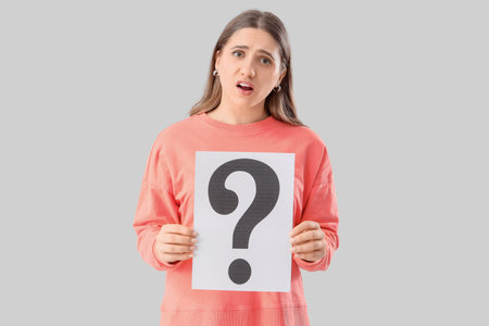 Puzzled woman holding paper sheet with question mark on gray background