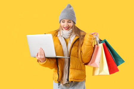 Beautiful young woman in winter clothes with laptop and shopping bags on yellow backgroundの写真素材
