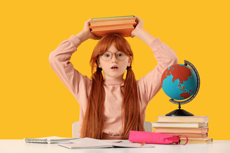 Shocked teenage redhead girl with books at table on yellow background