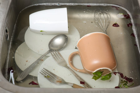 Metallic kitchen sink with soap water, dirty dishes and food leftovers, closeup