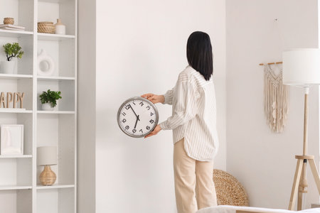 Young woman hanging clock on light wall at home, back view
