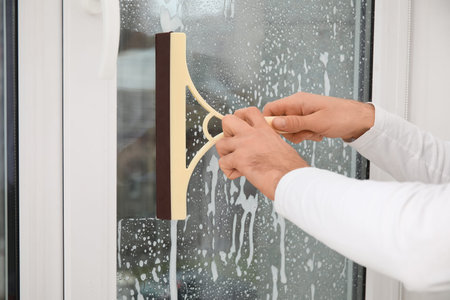 Male worker with squeegee washing window in room, closeup