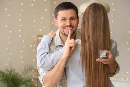 Young man with engagement ring showing silence gesture and hugging his girlfriend at homeの写真素材