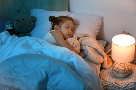 Cute little girl with knitted toy bunny sleeping in bedroom at nightの写真素材