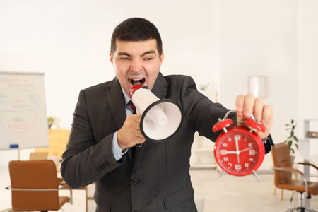 Young businessman with alarm clock shouting into megaphone in office. Time management conceptの写真素材