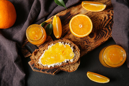 Wooden board with slices of bread and jar of sweet orange jam on black backgroundの写真素材