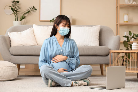 Pregnant Asian woman in medical mask with laptop sitting on floor at home