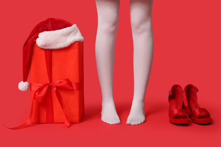 Legs of young woman in white tights with stylish high heels, gift box and Santa hat on red background. Christmas celebrationの写真素材
