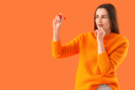 Wondering young woman with marker writing on orange backgroundの写真素材