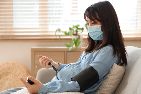 Pregnant Asian woman in medical mask measuring blood pressure on sofa at home