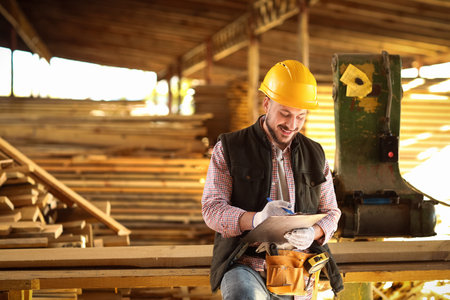 Young carpenter in hardhat with clipboard working at sawmill