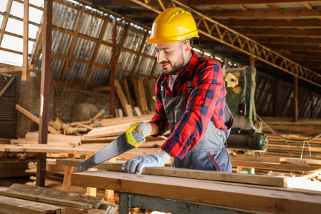 Young carpenter in hardhat sawing wooden plank at sawmill