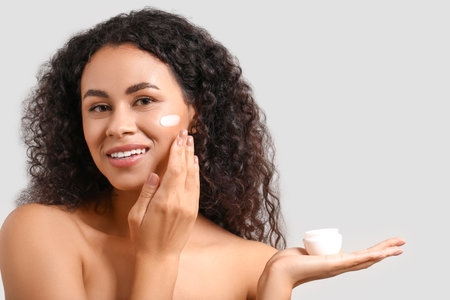 Beautiful young happy African-American woman with healthy skin and jar of cream on gray backgroundの写真素材