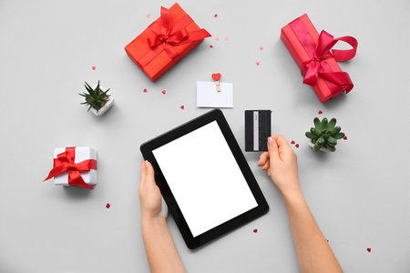 Female hands with modern tablet computer, credit card and gifts for Valentine's Day on gray backgroundの写真素材