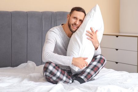 Young man in pajamas with soft pillow sitting on bed in bedroom. World Sleep Dayの写真素材