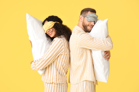 Beautiful young couple in pajamas and masks with soft pillows on yellow background. World Sleep Dayの写真素材