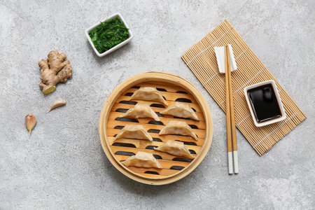 Bamboo steamer with tasty Japanese gyoza and bowls of soy sauce and seaweed salad on gray background