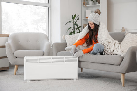 Frozen young woman with plaid sitting on sofa near electric convector heater at homeの写真素材