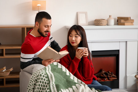Loving young happy couple with cup of tea reading book on sofa near fireplace at homeの写真素材
