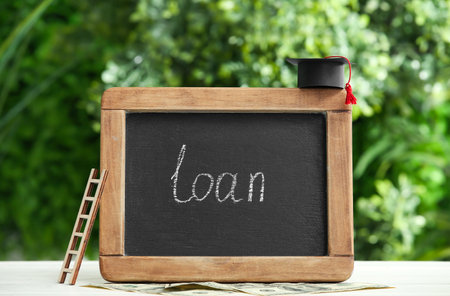 Chalkboard with word LOAN, graduation hat, ladder and money on table outdoorsの写真素材